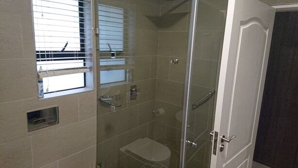 A glass-enclosed shower stall with a toilet, a window with blinds, and a door with a handle. - Unitas Guesthouse (Centurion)