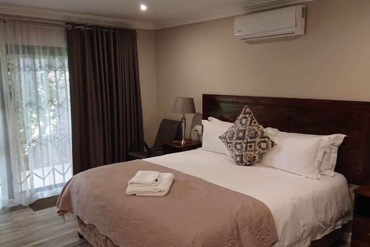 Una habitación de hotel con una cama grande, una mesita de noche, una lámpara, un equipo de aire acondicionado y vistas al exterior a través de una ventana con cortinas.