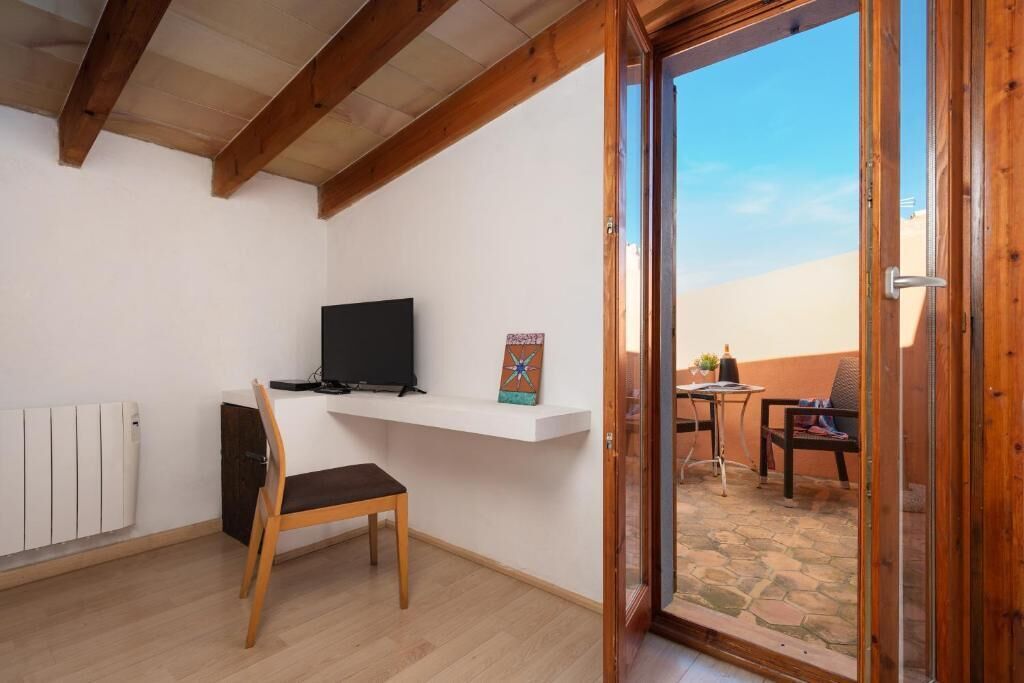 A room with a wooden ceiling, a white wall, a TV on a shelf, a chair, and a door leading to an outdoor patio with a table and chairs.