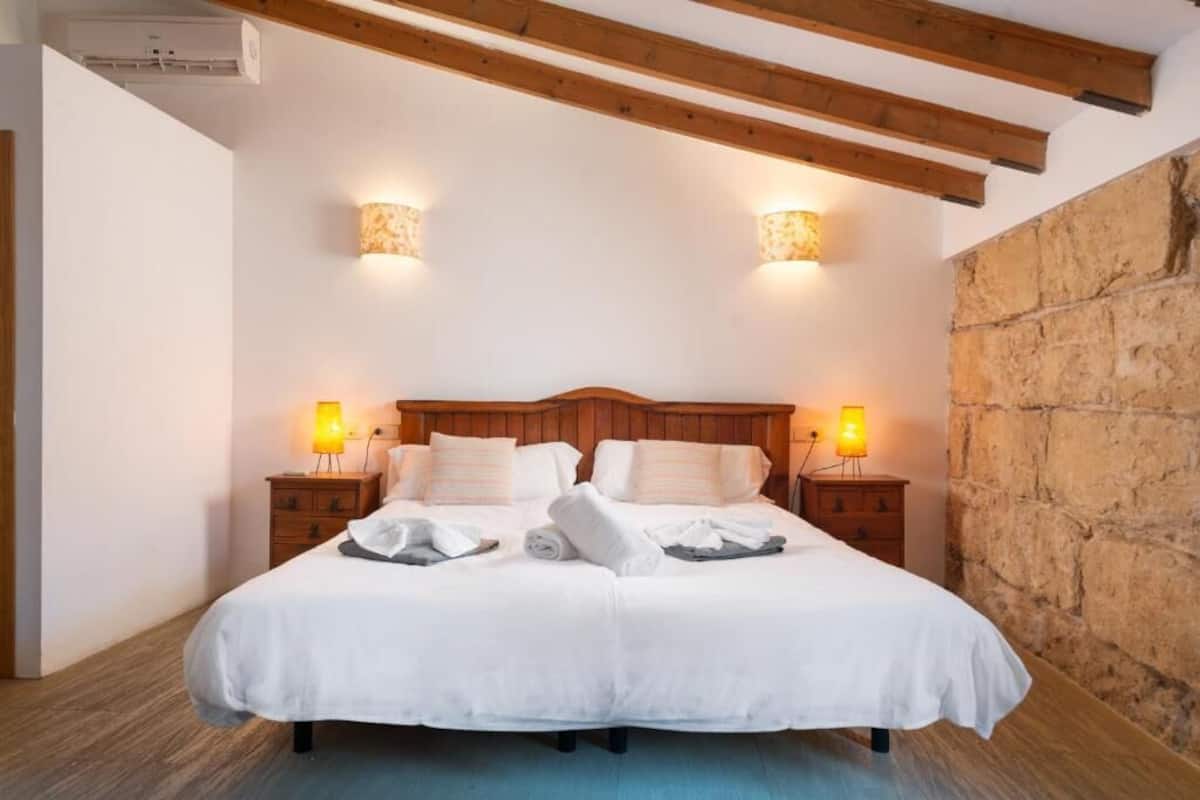 Cama doble con sábanas blancas, cabecera de madera, dos lámparas de noche y pared de piedra.