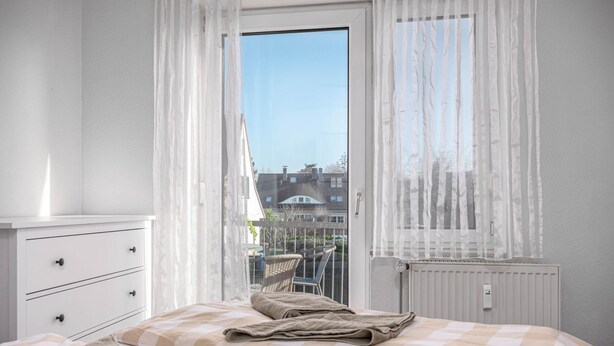 A bedroom with a bed, a nightstand, a window with sheer curtains, and a view of buildings outside. - Bahnhofstraße 10 - Apartment 3 - Holiday apartment in Zingst with balcony near the beach (Zingst)