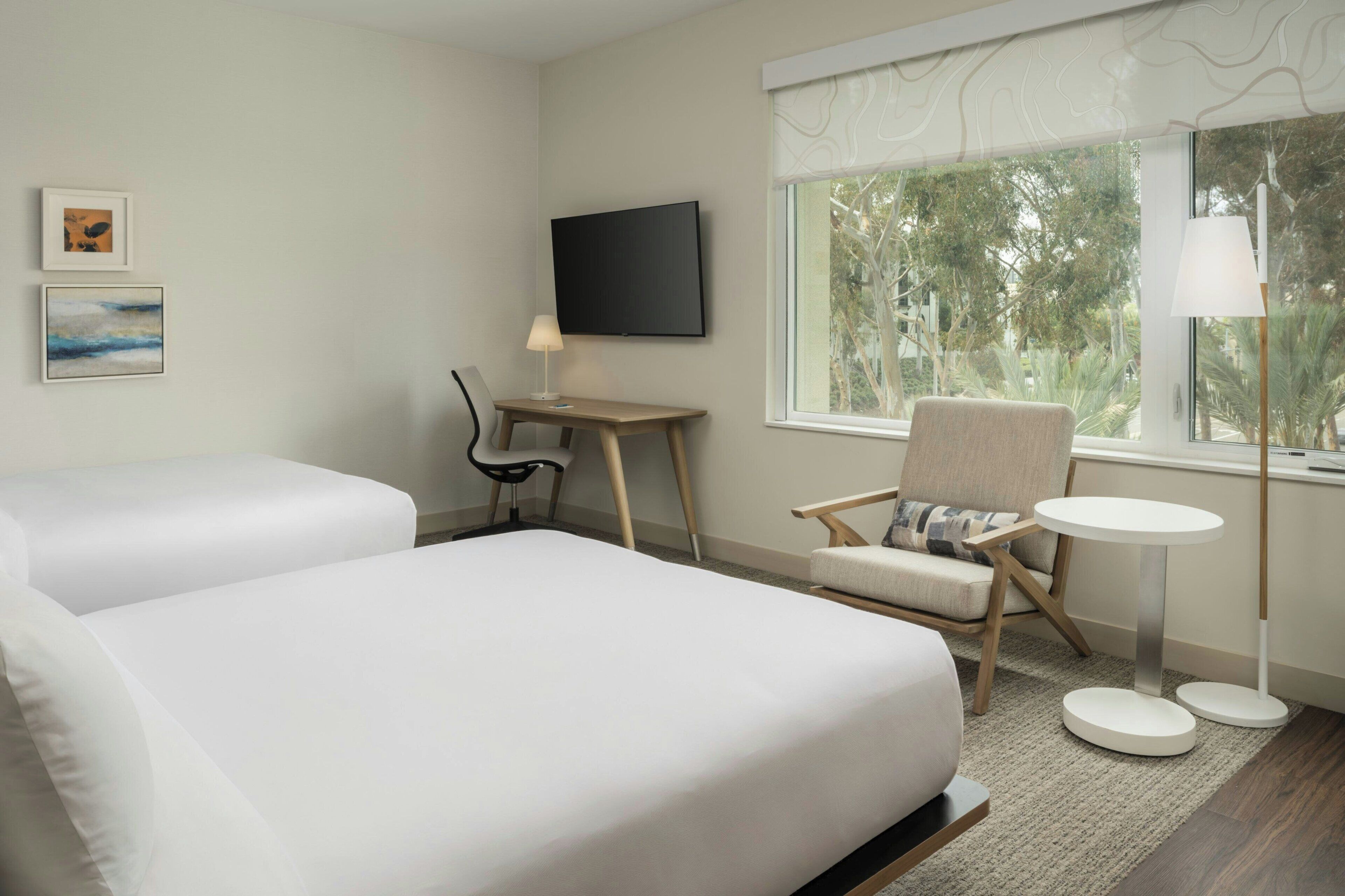 A hotel room with two beds, a desk, a chair, a TV, and a window with a view of trees.