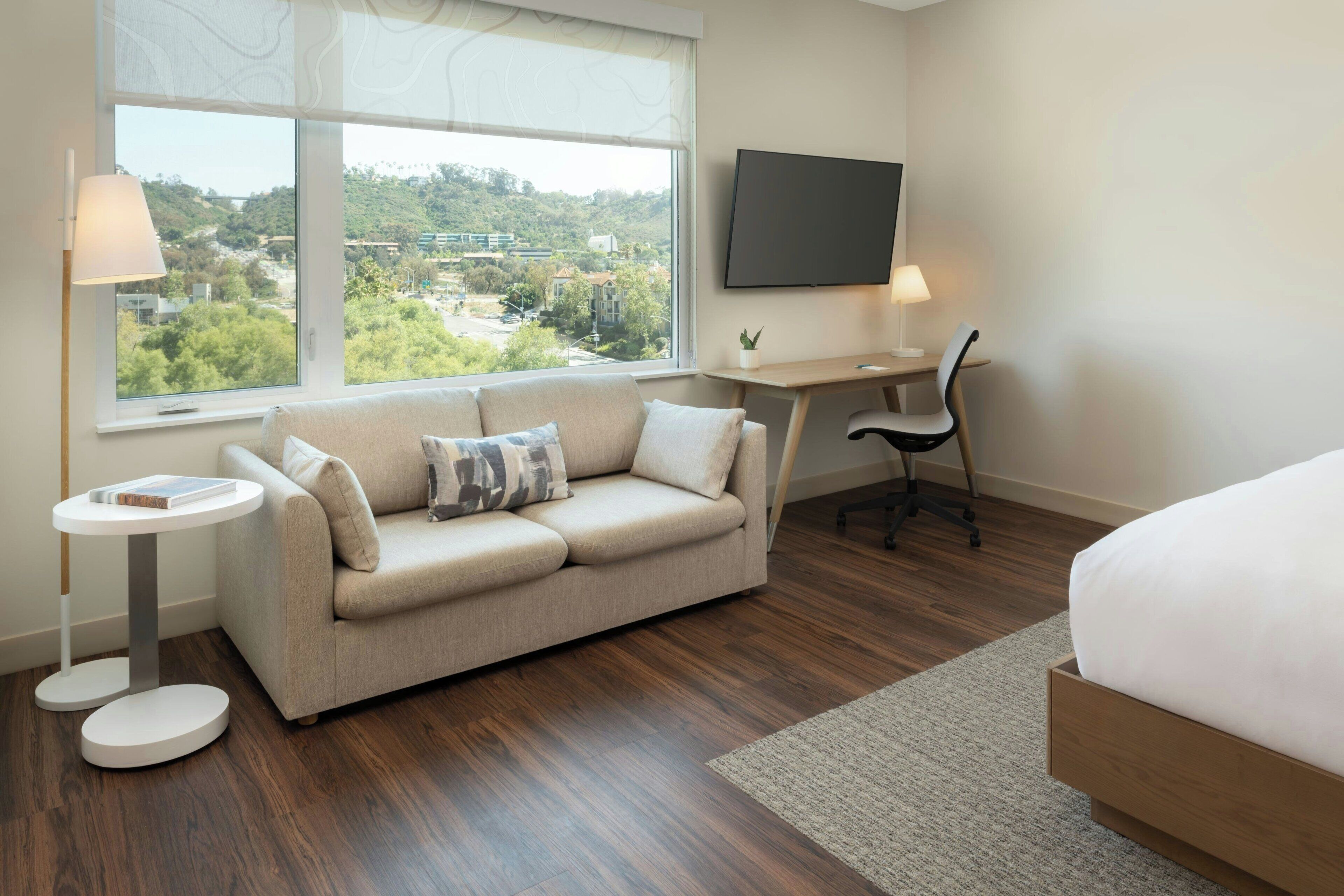 A modern hotel room with a sofa, a desk, a chair, a bed, and a TV.