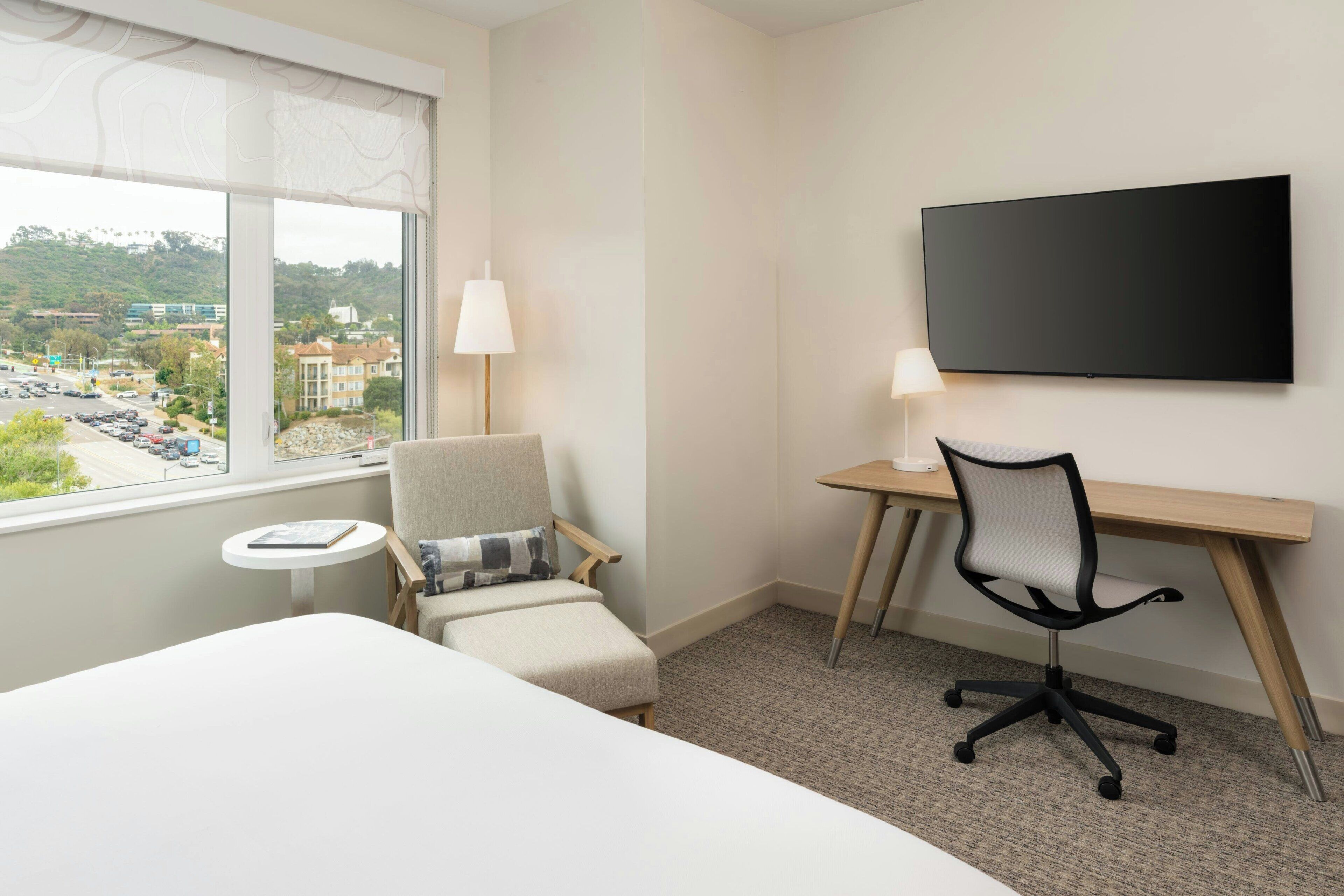 A hotel room with a bed, a desk, a chair, a TV, and a view of a cityscape.