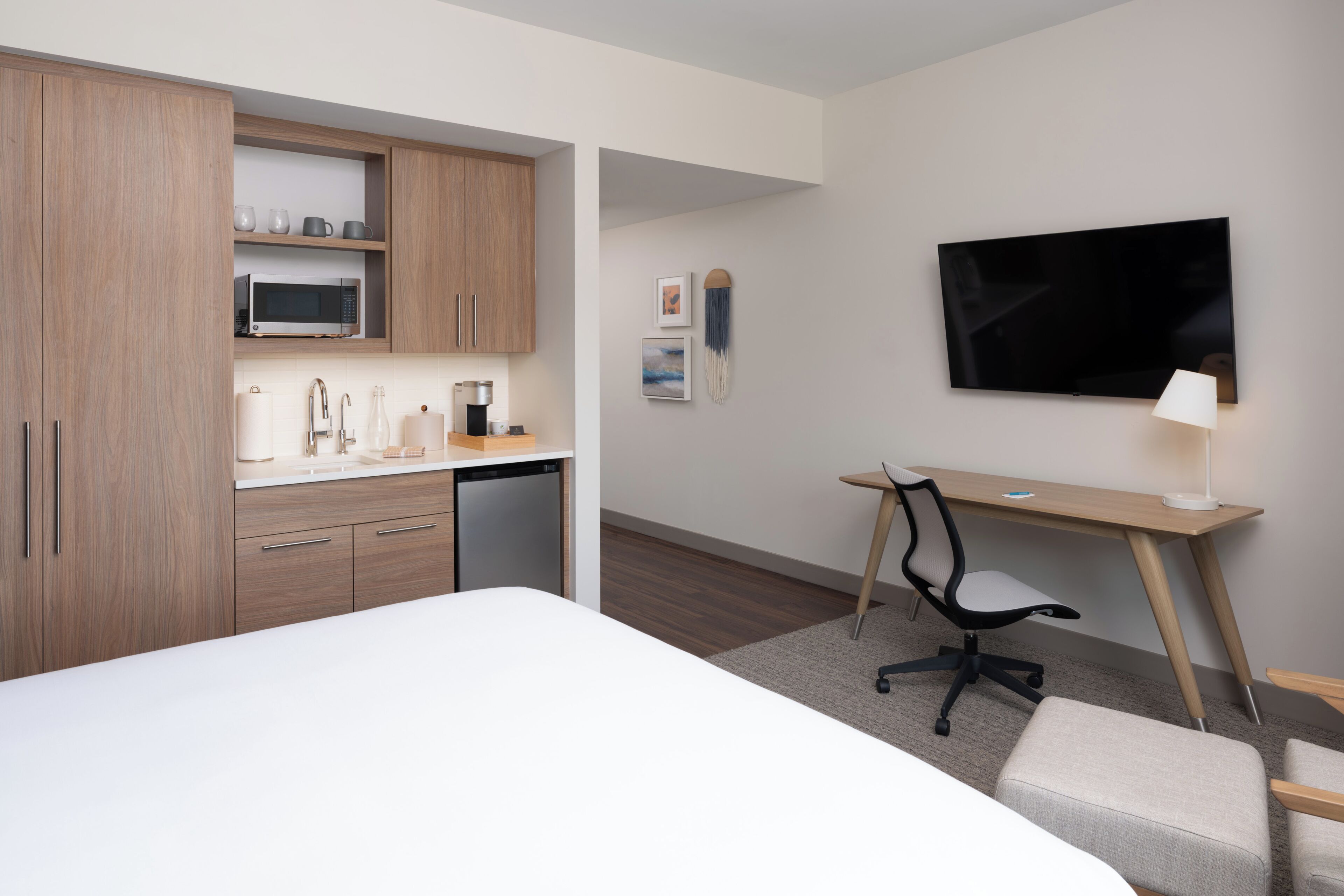 A hotel room with a bed, a desk with a chair, a television, a microwave, and a small kitchenette.