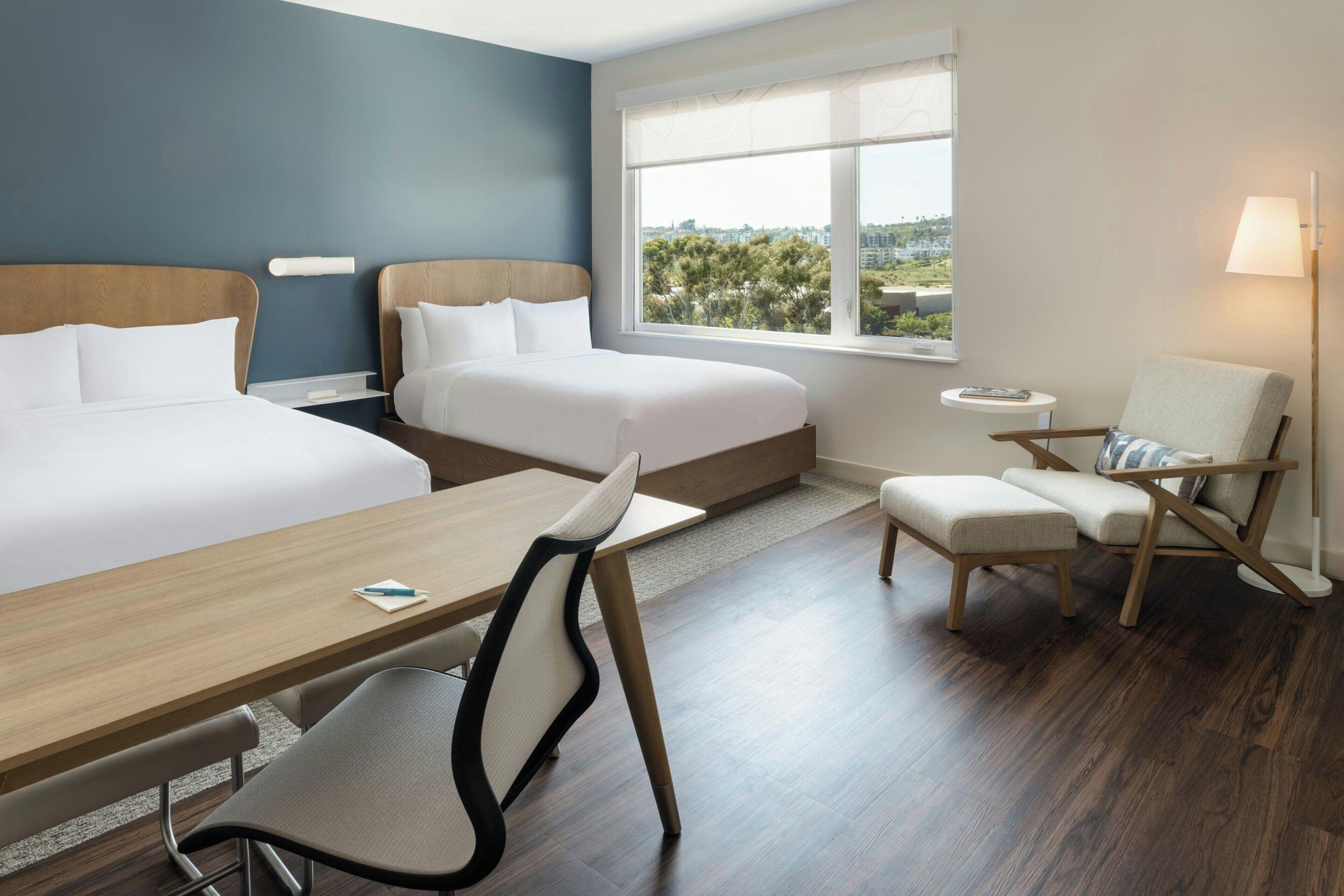 A hotel room with two beds, a desk, a chair, a lamp, and a view of the outdoors.