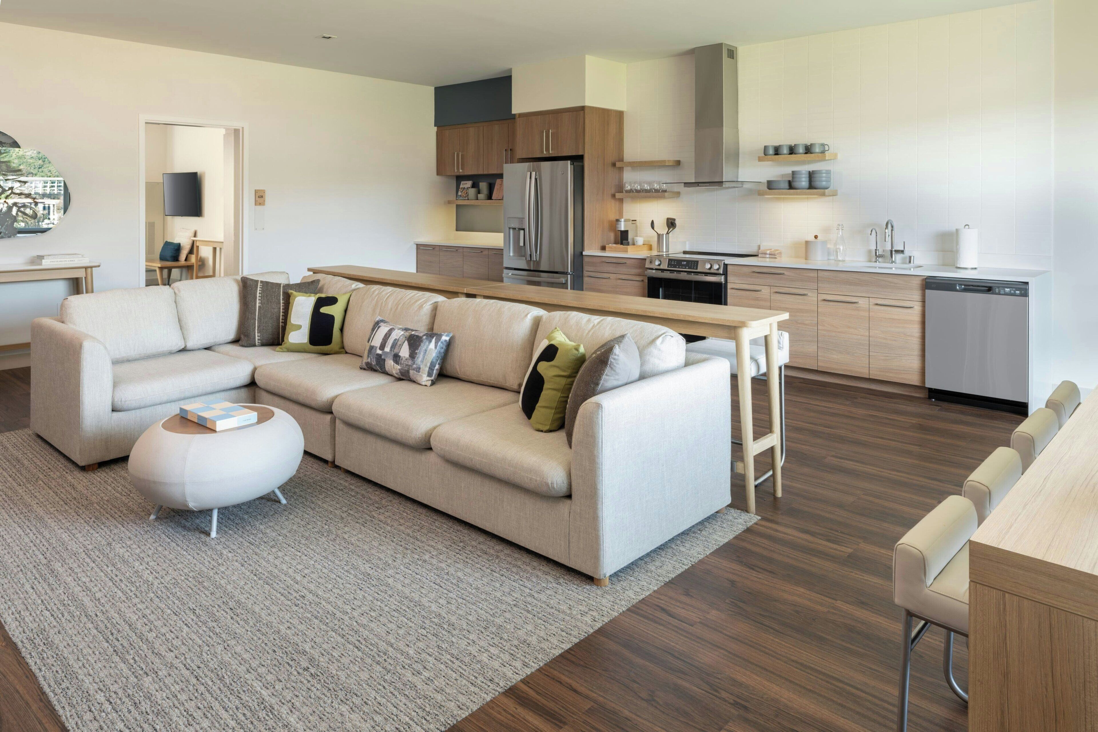 Un salon moderne avec un canapé d’angle, un coin repas doté d’une table en bois et une cuisine équipée d’appareils électroménagers en acier inoxydable.