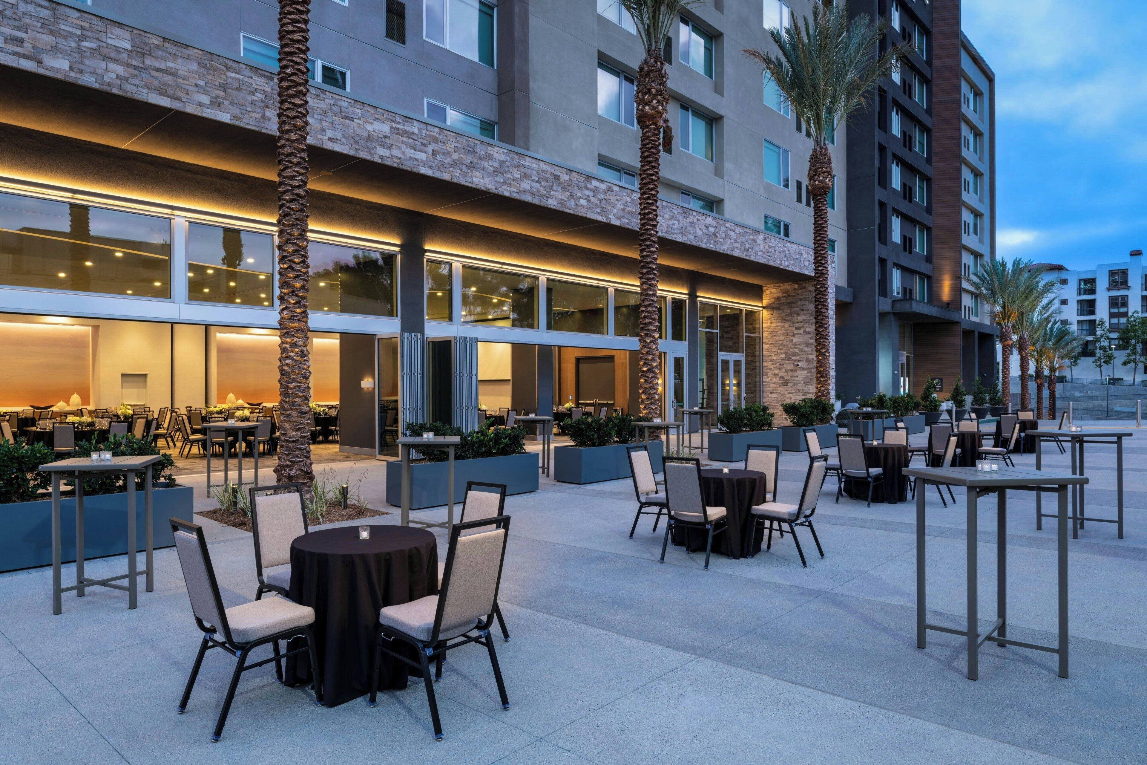 An outdoor dining area with tables and chairs, a building with large windows, and palm trees.