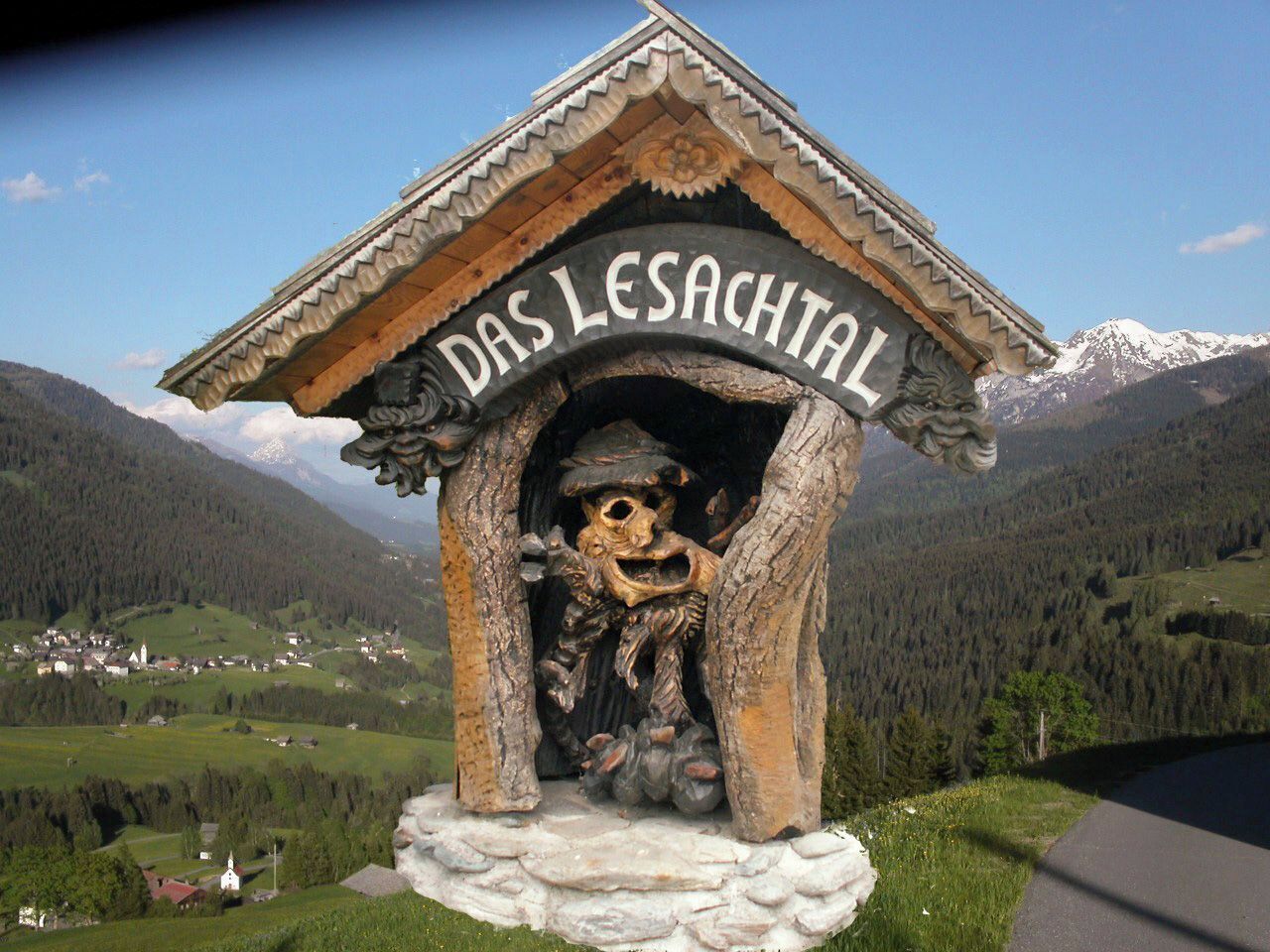 Une structure en bois portant l’inscription « DAS LESACHTAL » et ornée d’une sculpture, le tout se dressant sur un fond de paysage montagneux.