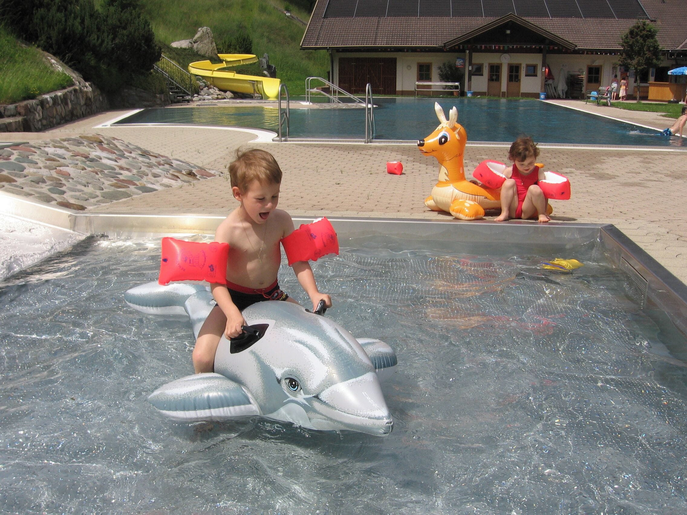 A pool area with inflatable toys, a building with a tiled roof, and a stone wall.