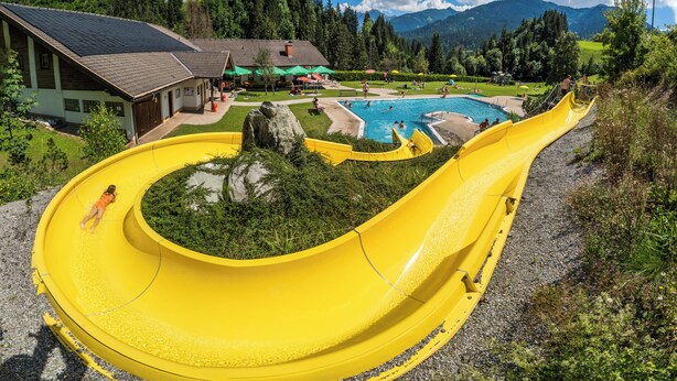Un tobogán acuático amarillo que desemboca en una piscina, rodeado de vegetación y con una montaña al fondo.