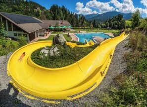 Un toboggan aquatique jaune se terminant dans une piscine, entouré de verdure et avec une montagne en toile de fond.