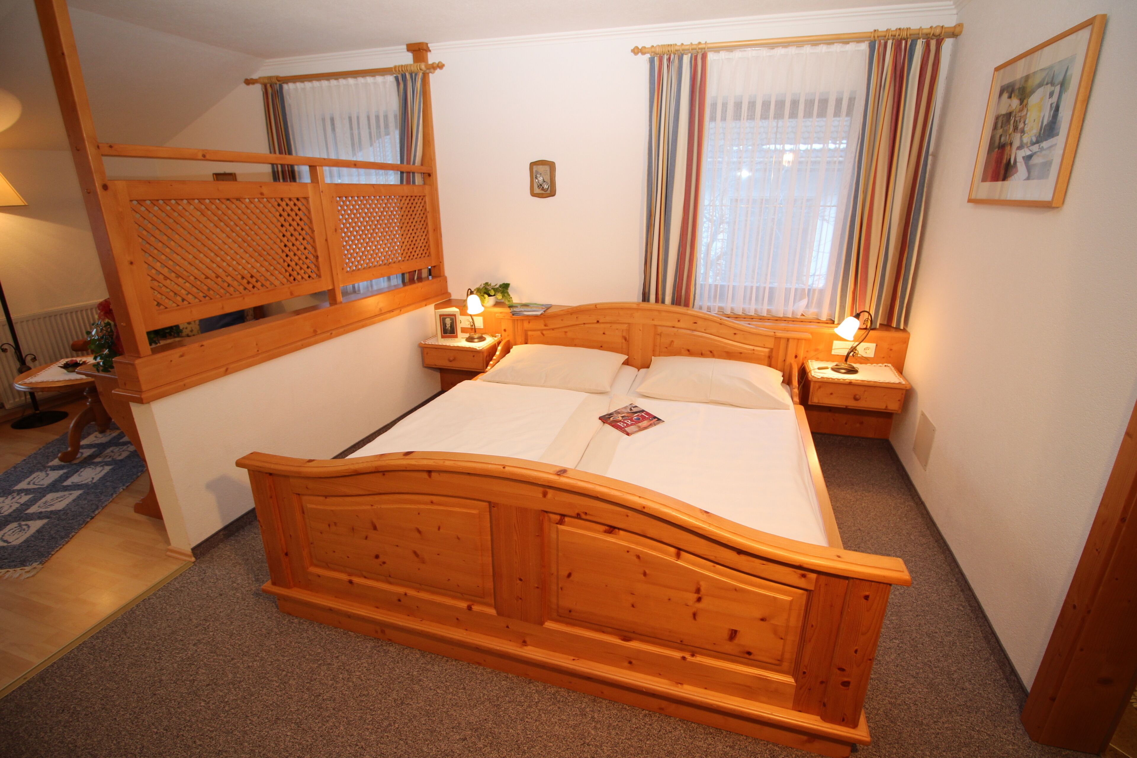 Una cama de madera con cabecera, dos mesitas de noche con lámparas, una ventana con cortinas, y un cuadro enmarcado en la pared.