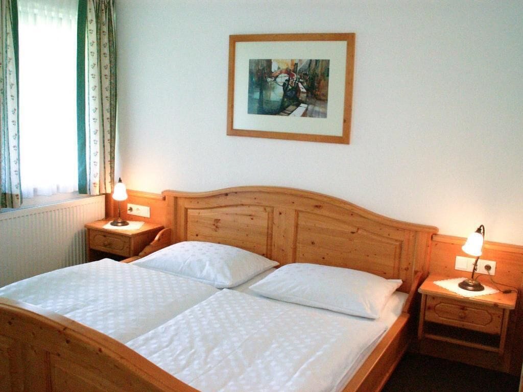 A double bed with two pillows, wooden headboard, bedside tables with lamps, a framed picture on the wall, and a window with curtains.