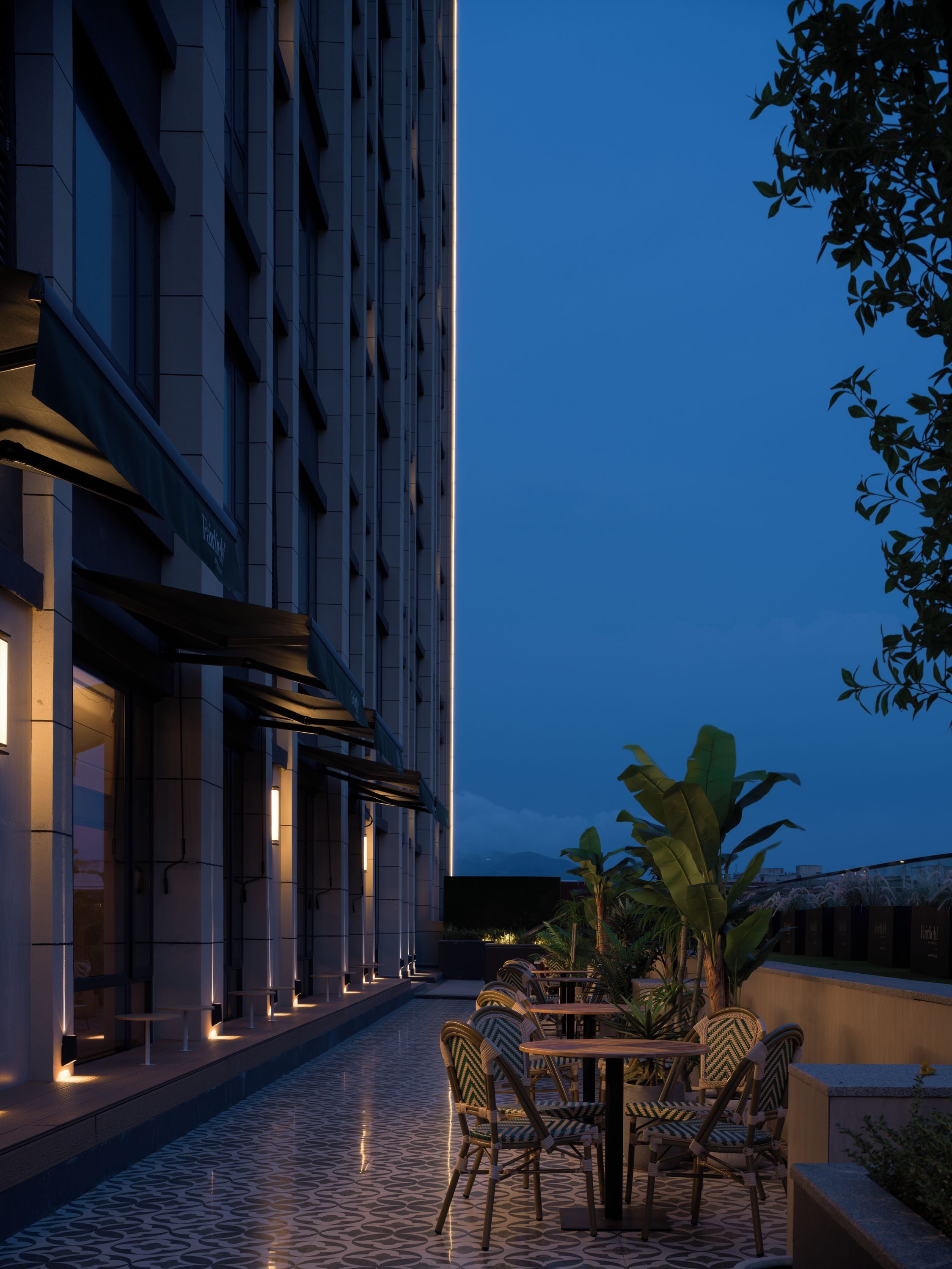 A modern outdoor seating area with tables and chairs, a tiled floor, and a building facade with illuminated windows.