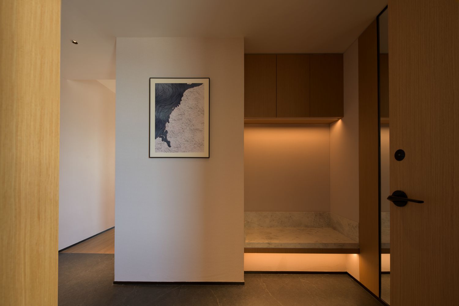 Un couloir orné d’un tableau encadré au mur, une porte en bois et un banc avec un éclairage intégré en dessous.