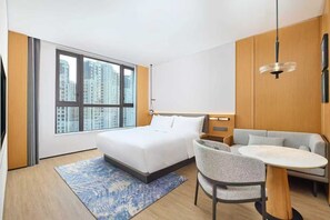 Habitación de hotel moderna con una cama grande, un sofá, una mesa de comedor y vistas a edificios altos a través de amplias ventanas.