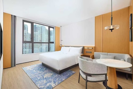 A modern hotel room with a large bed, a sofa, a dining table, and a view of tall buildings through large windows.