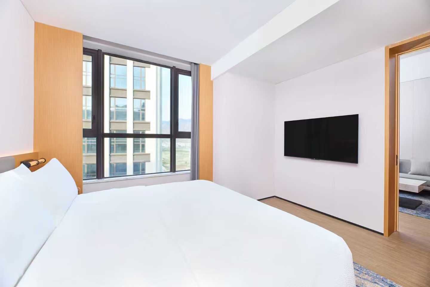 A modern hotel room with a large bed, a TV mounted on the wall, and a window with a city view.
