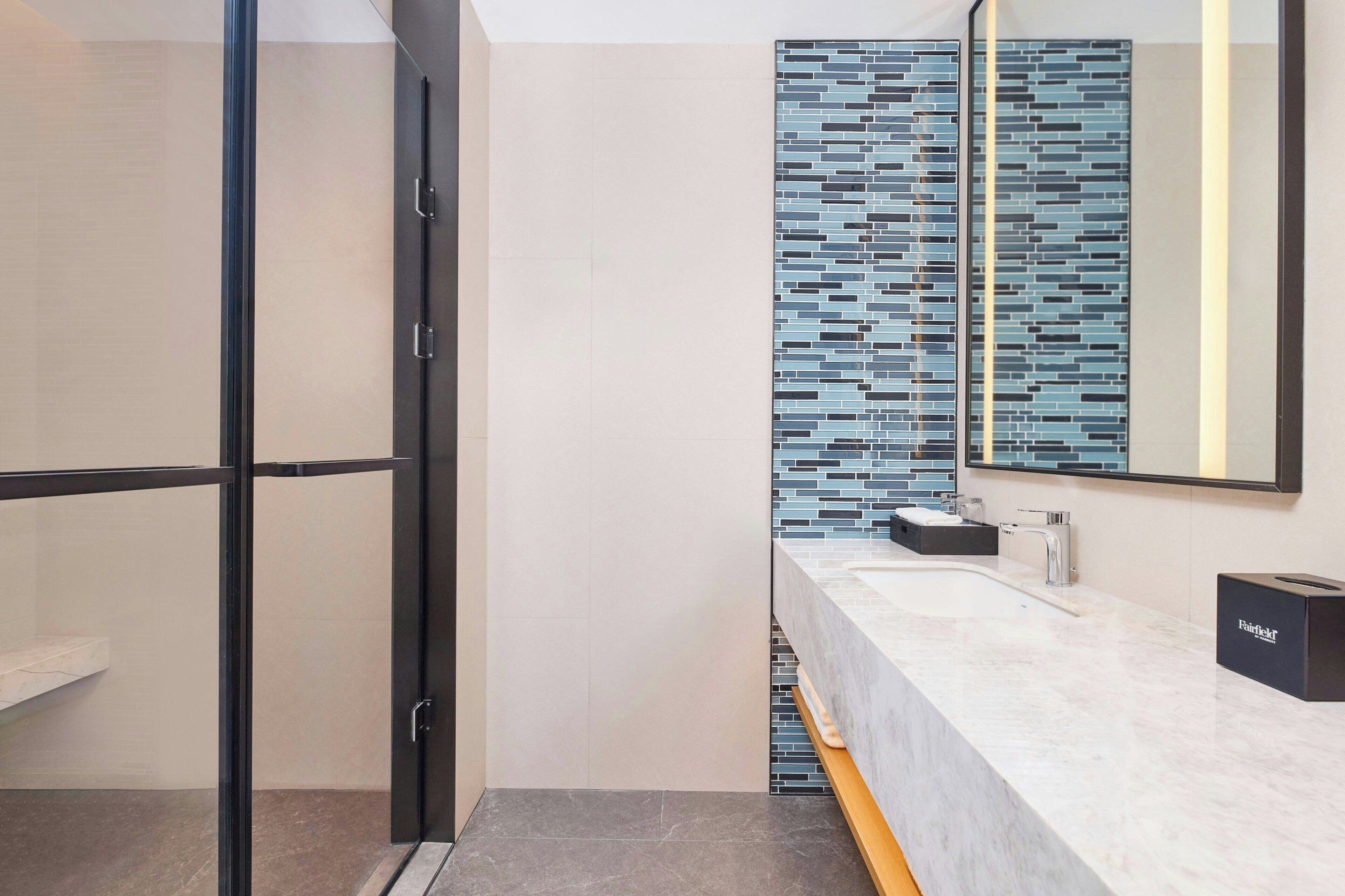 A modern bathroom with a glass shower enclosure, a tiled backsplash, and a marble countertop with a sink and a towel dispenser.