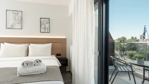 A hotel room with a bed, a nightstand, a window with curtains, and a balcony with a table and chair. - Tower 15 | Deluxe Studio 402 (Thessaloniki)