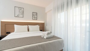 A modern hotel room with a large bed, a headboard, two framed artworks, a neatly made bed with towels, and sheer curtains. - Tower 15 | Deluxe Studio 402 (Thessaloniki)