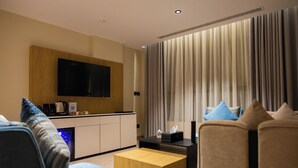 A modern hotel room with a flat-screen TV mounted on a wooden panel, a kitchenette, a seating area with a sofa and armchairs, and a large window with curtains. - Lamar Park Hotel Al Olaya (Tabuk)