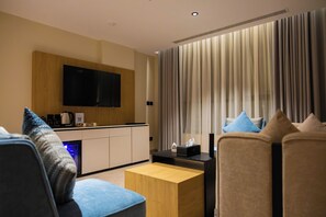 A modern hotel room with a flat-screen TV mounted on a wooden panel, a kitchenette, a seating area with a sofa and armchairs, and a large window with curtains. - Lamar Park Hotel Al Olaya (Tabuk)