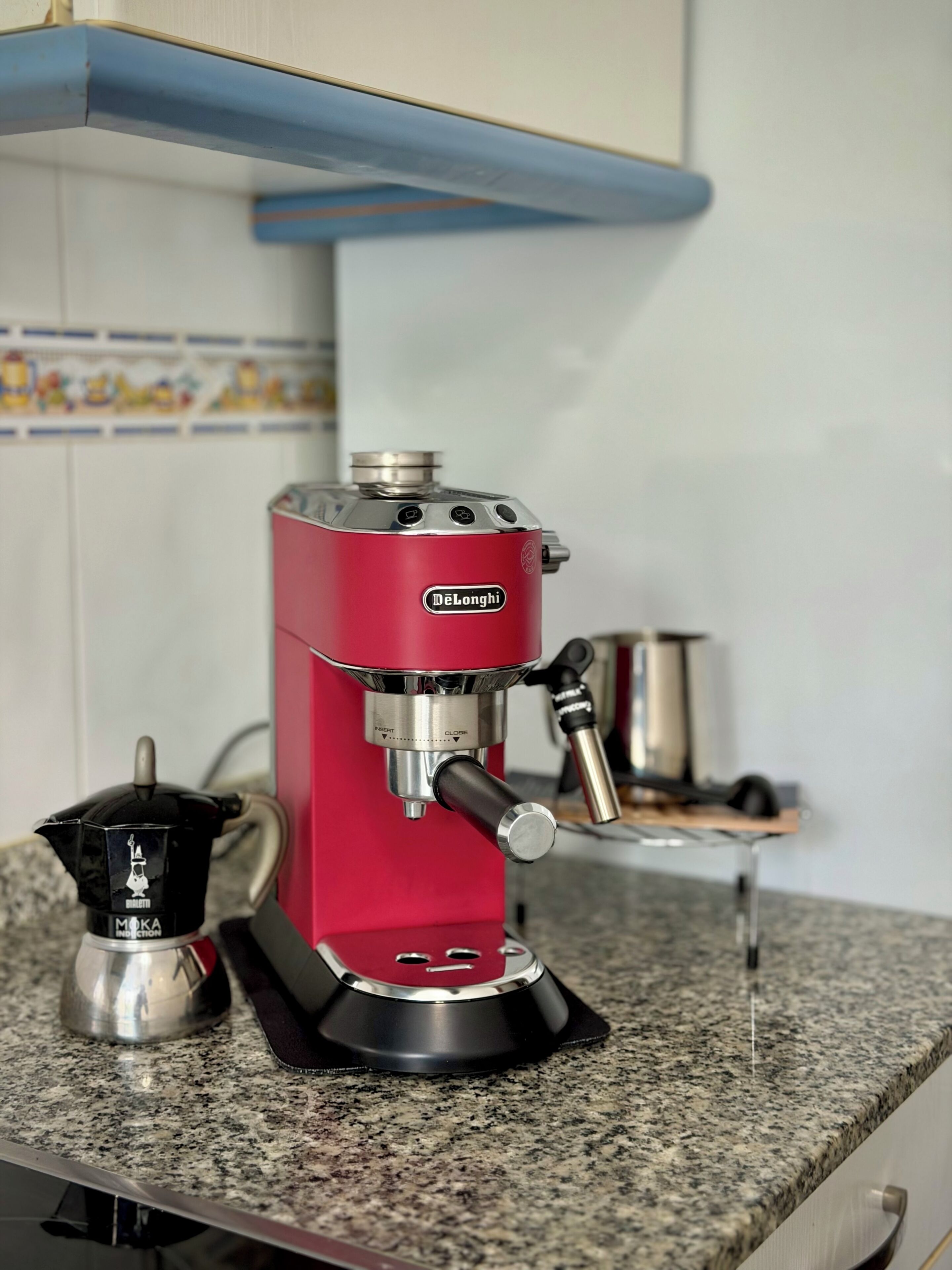 A red espresso machine on a granite countertop with a coffee pot and a small metal container.