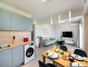 A modern kitchen with a washing machine, a dining area with breakfast setup, a grey sofa, and a flat-screen TV. - Asmira Apartments (Lefkada)