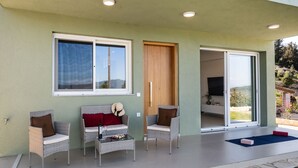 Ein moderner Außenbereich mit Korbstühlen, einem Glastisch und Blick auf eine gebirgige Landschaft durch große Fenster.