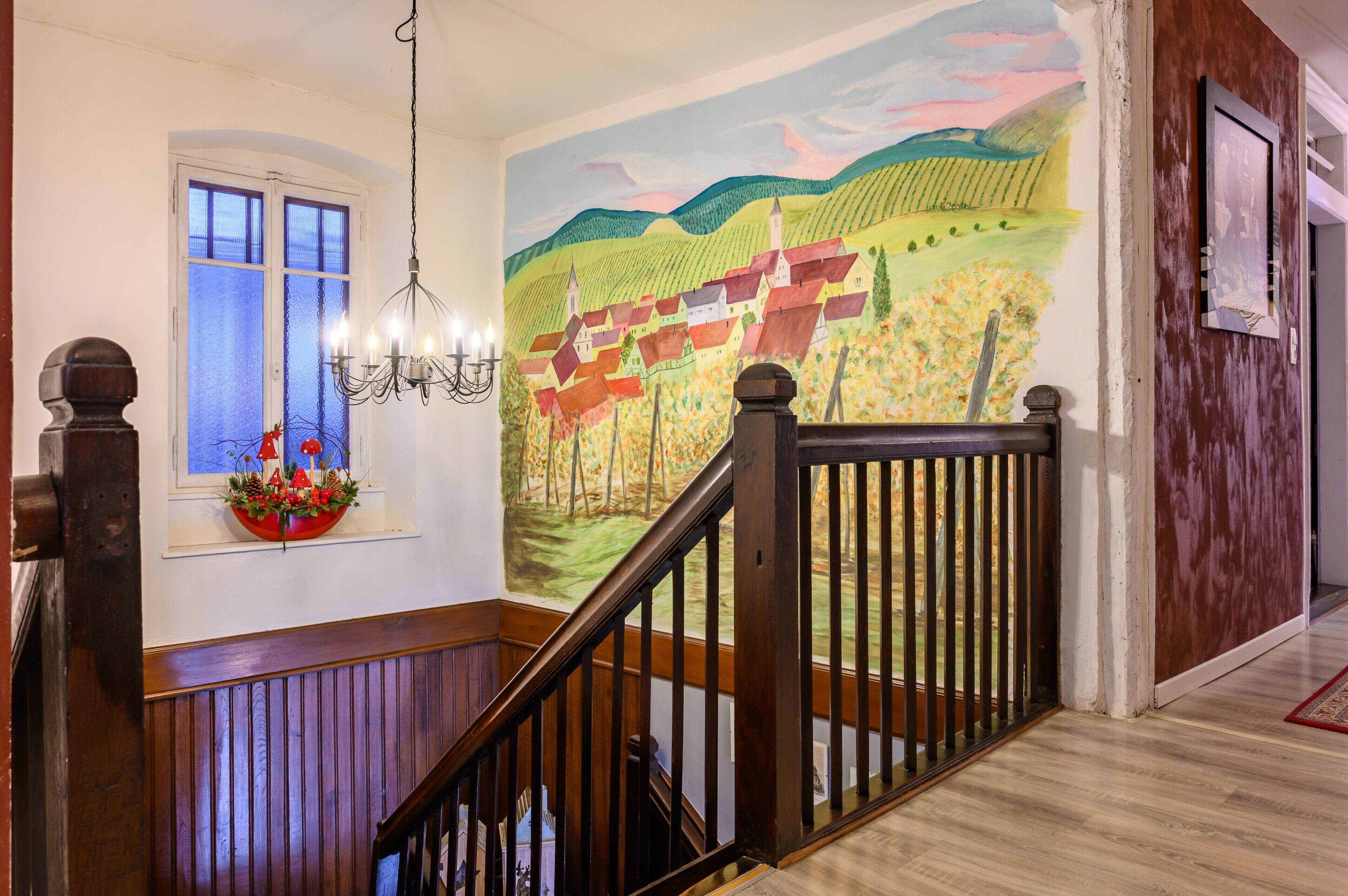 Una escalera con barandilla de madera y pasamanos, un candelabro, una ventana con una cesta de flores y un mural en la pared.