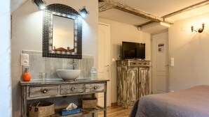 A bathroom with a large mirror, sink, and vanity with storage. There's a television on a wooden cabinet and a bed with a patterned bedspread. - Bed and Breakfast 'Demeure D'antan - Chambre Esmeralda' with Shared Terrace, Shared Garden and Wi-Fi (Beblenheim)