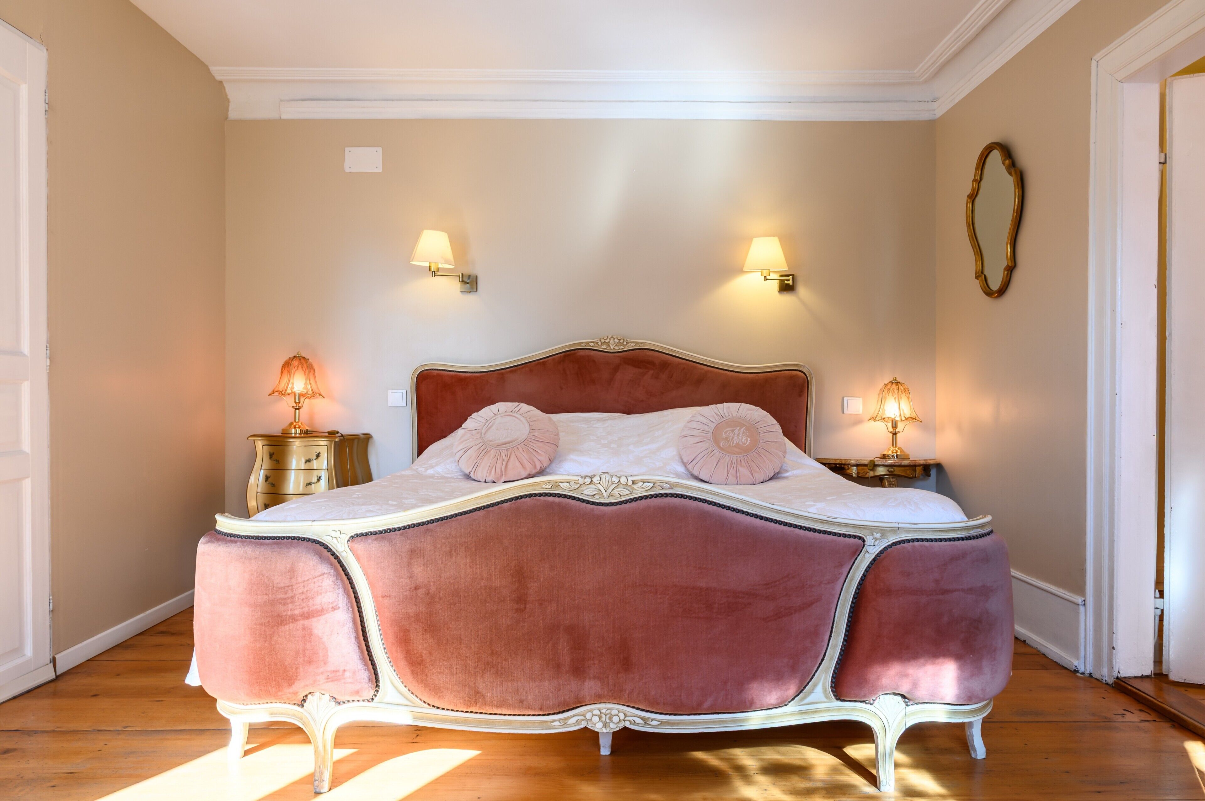 Een bed met een zacht, rood hoofdeinde, twee bedlampen en een spiegel aan de muur.