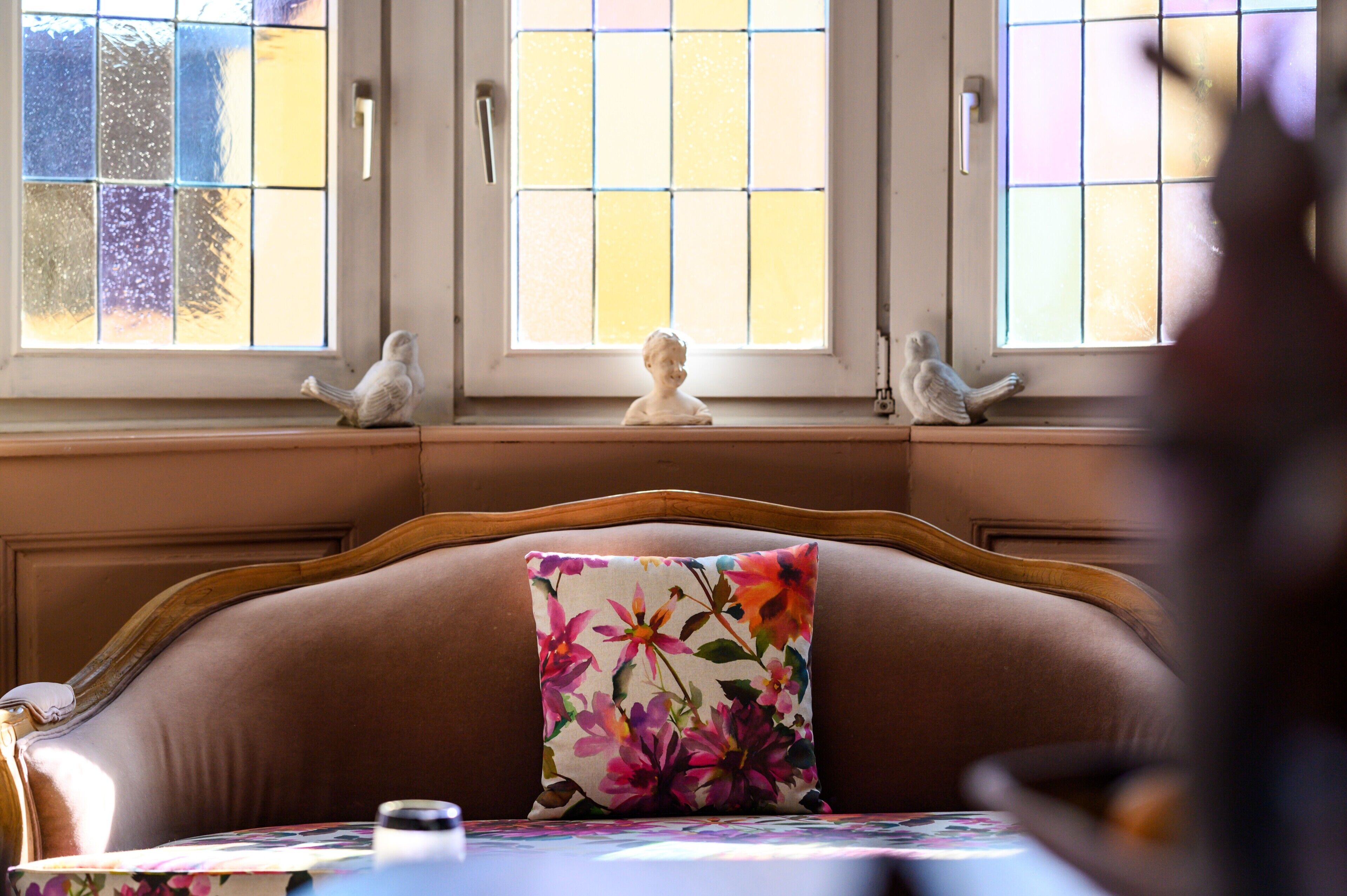 Een kamer met een comfortabele bank, een bloemenkussen en twee vogelbeeldjes op de vensterbank.