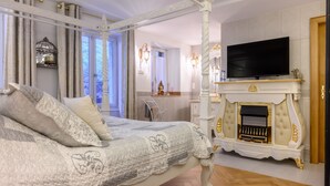 Ein Bett mit Baldachin, eine Patchworkdecke, ein Kamin mit Fernseher darüber und ein Spiegel mit Wandleuchten.