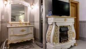 Ein luxuriöses Badezimmer mit einem großen Spiegel, einer aufwendigen gold- und weißfarbenen Waschtischgarnitur und einem Fernseher über einem Kamin.
