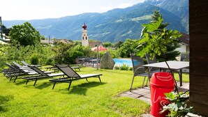 Eine Terrasse mit Liegestühlen, einem roten Mülleimer und Blick auf Berge sowie eine Kirche.