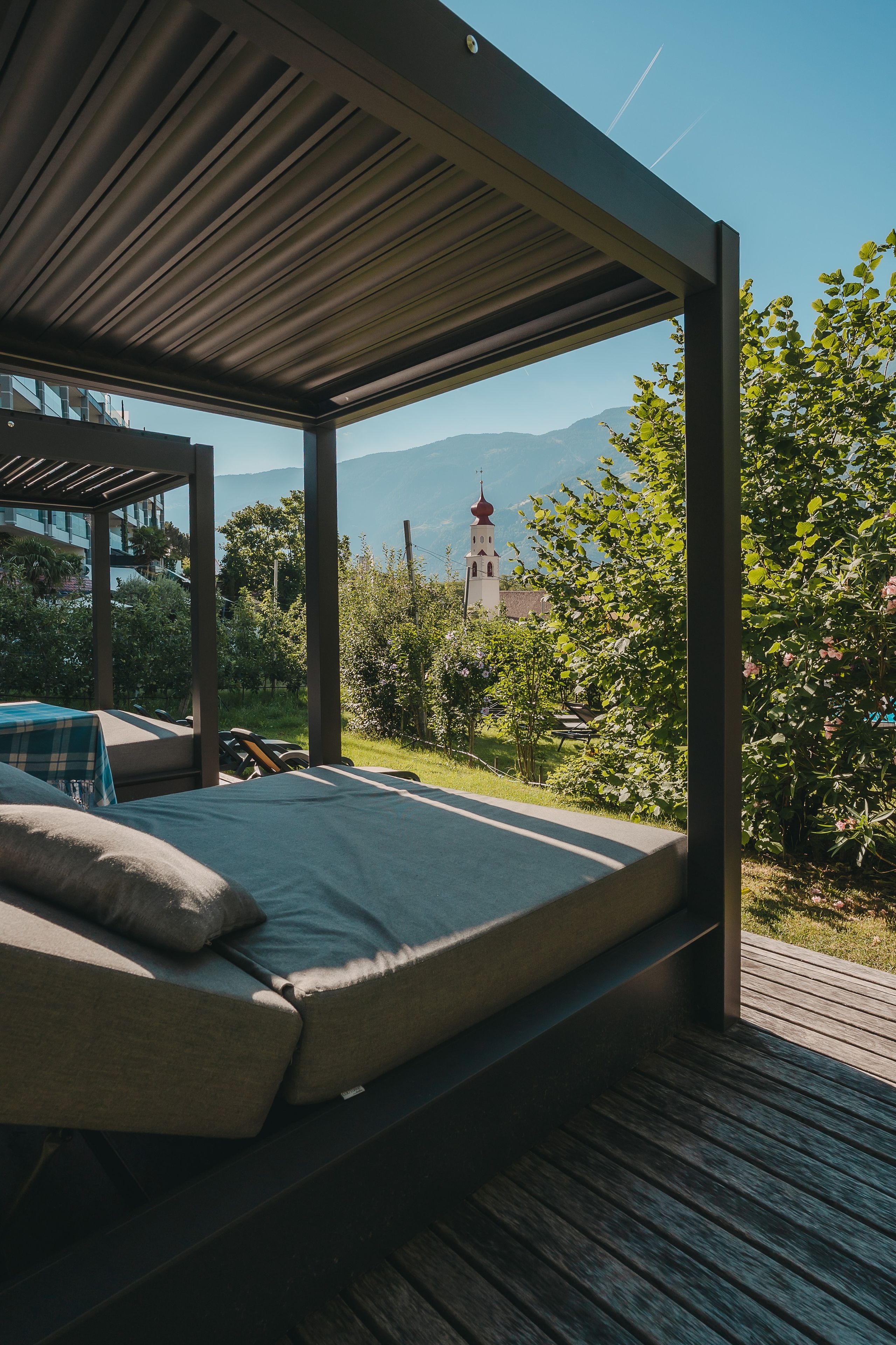 A bed in a covered outdoor area with a view of a church and mountains.