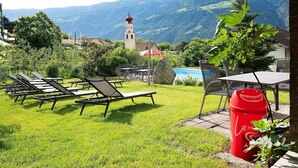 Ein grüner Außenbereich mit Liegestühlen, einem Tisch und einer roten Mülltonne.
