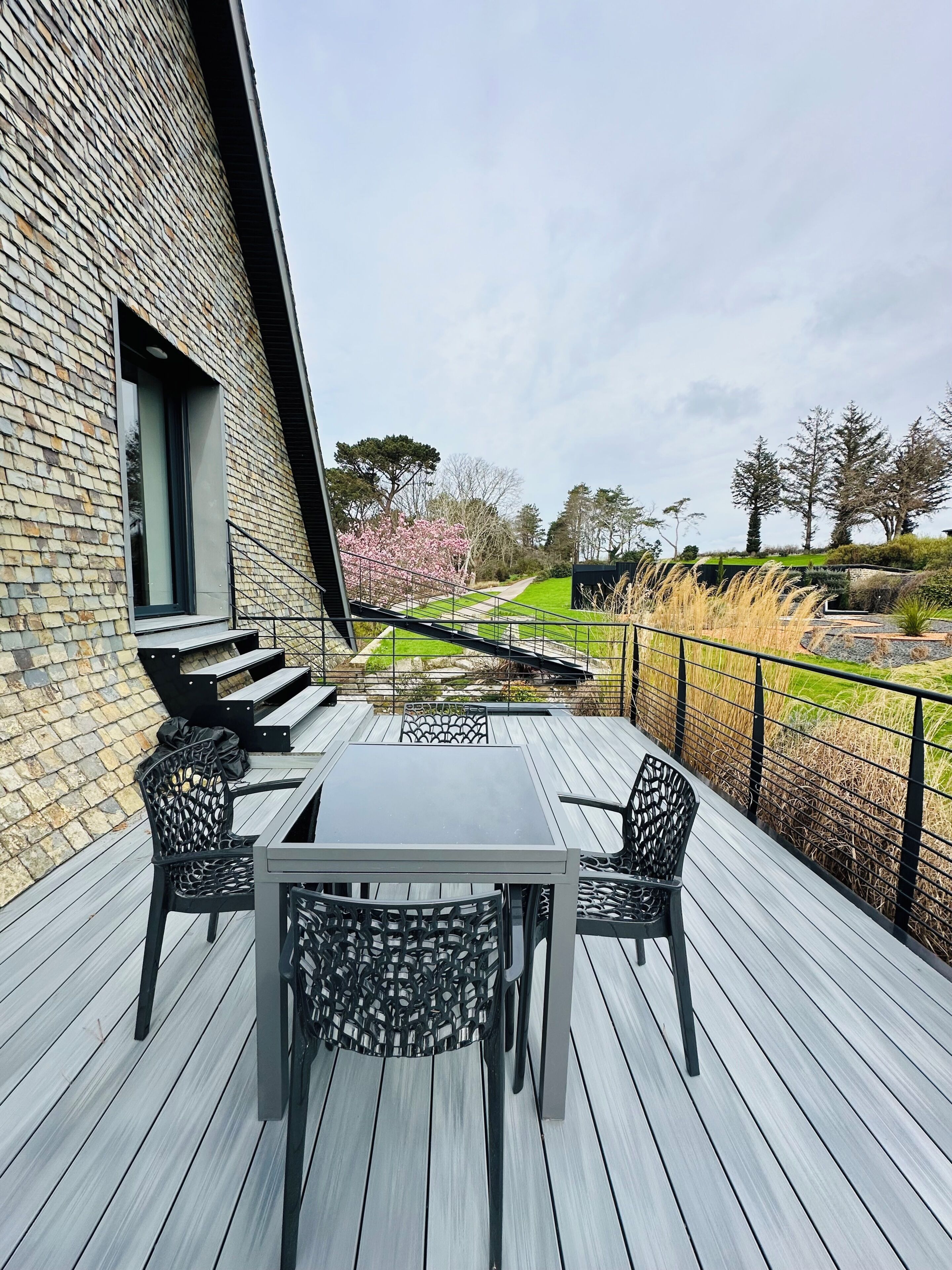 Eine Terrasse mit Tisch und Stühlen, eine Ziegelwand und Blick auf einen Garten mit Bäumen und einem Weg.