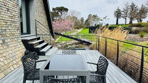 A deck with a table and chairs, a brick wall, and a view of a garden with trees and a path. - Avocet - Laïta (Guidel)
