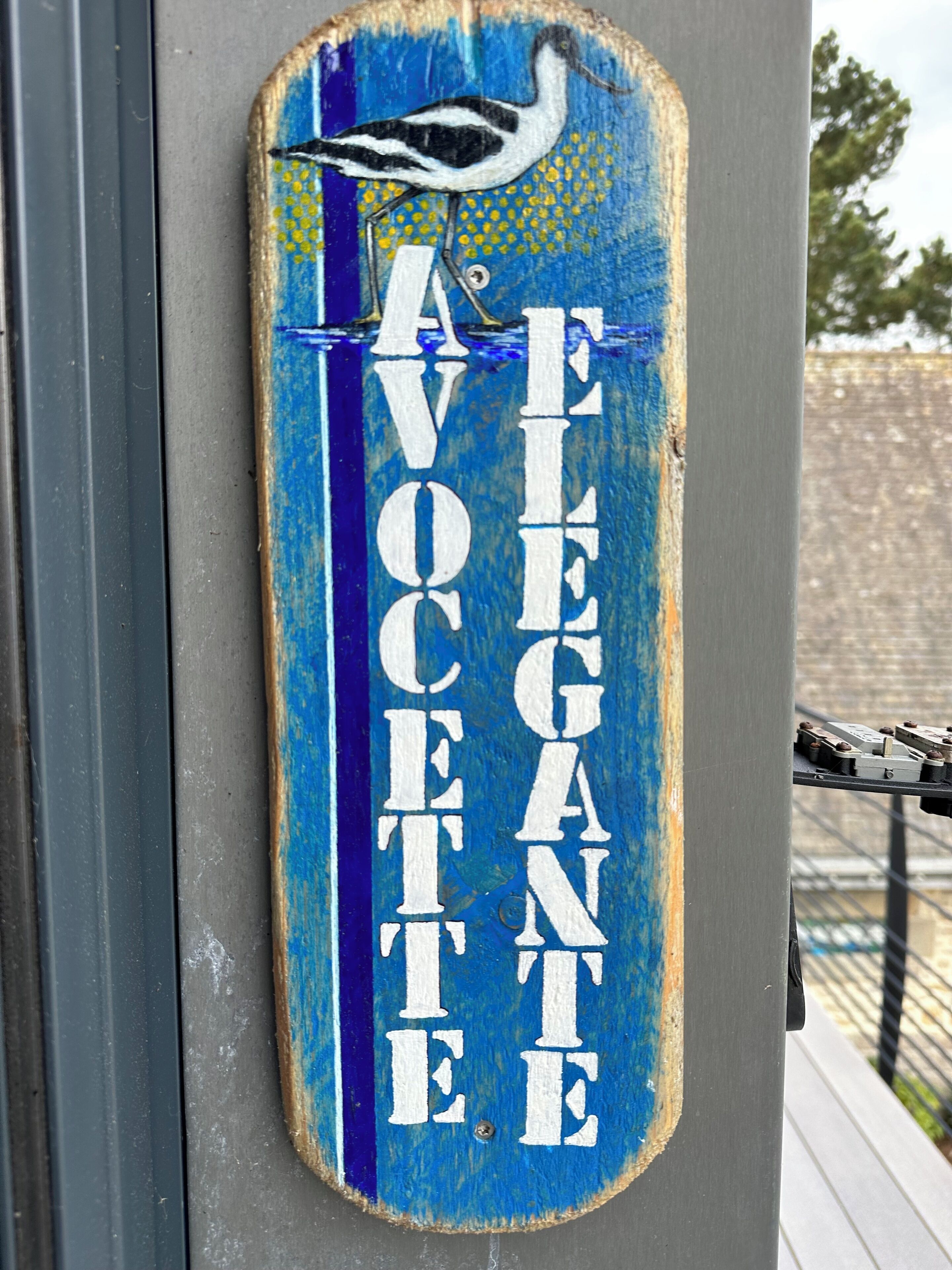 Un letrero azul con texto blanco e ilustración de un pájaro, sujeto a una estructura gris.