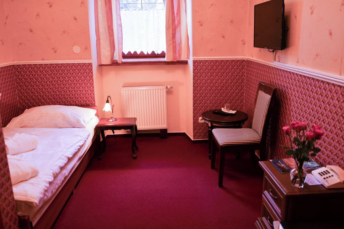 A room with a bed, a desk with a lamp, a chair, a radiator, and a television mounted on the wall.