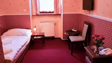 Una habitación con una cama, un escritorio con lámpara, una silla, un radiador y un televisor colocado en la pared.