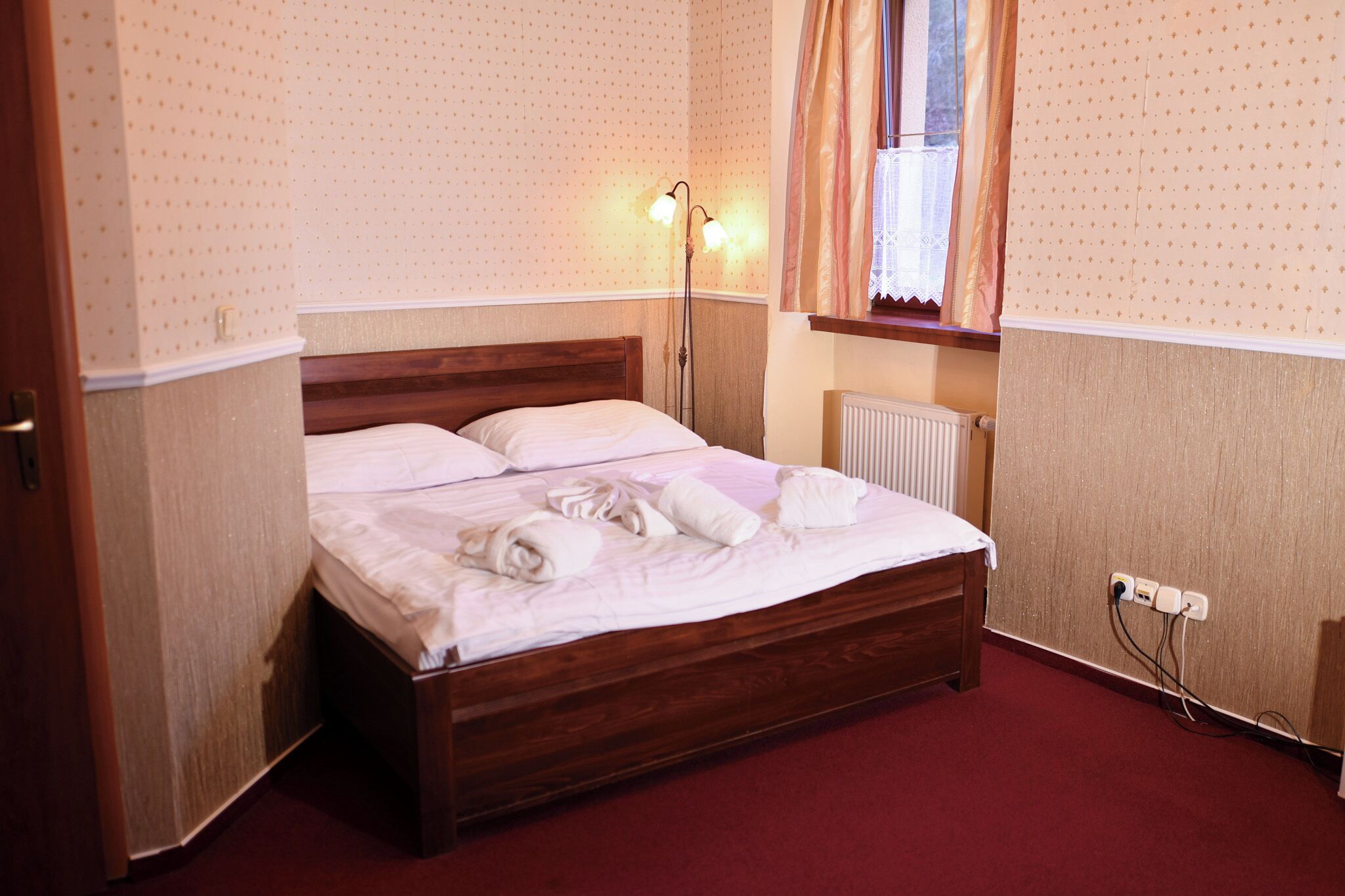 A neatly made bed with pillows and towels, a bedside lamp, a window with curtains, and a radiator.