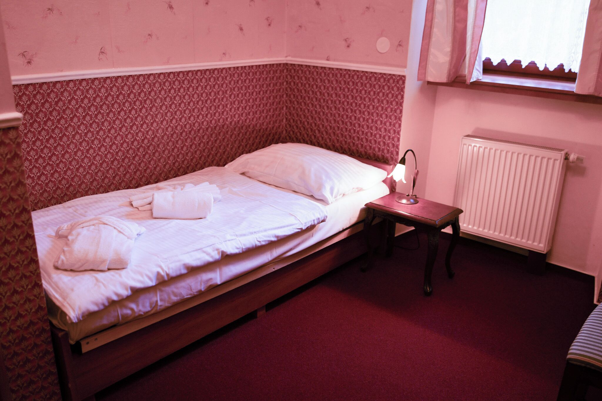 Una cama individual con estructura de madera, una mesita de noche con lámpara y un radiador en la pared.