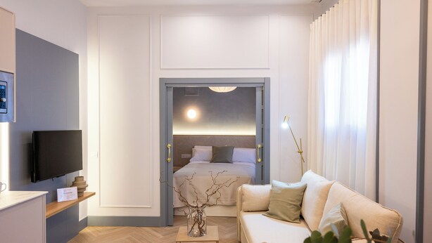 A modern hotel room with a flat-screen TV, a sofa, a bedside table, and a view into the bedroom.