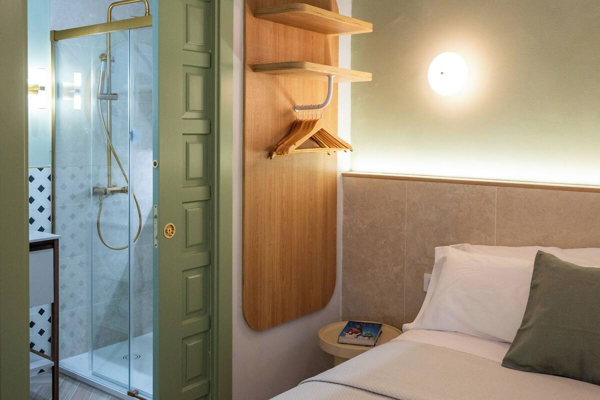 Ein Hotelzimmer mit Bett, Holzregal, Nachttisch, Wandlampe und Duschbereich.
