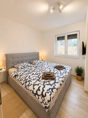 A bedroom with a large bed, a bedside lamp, a window with blinds, a wall-mounted TV, and a small plant. - Apartment Mariner 1 with heated pool (Zaton)